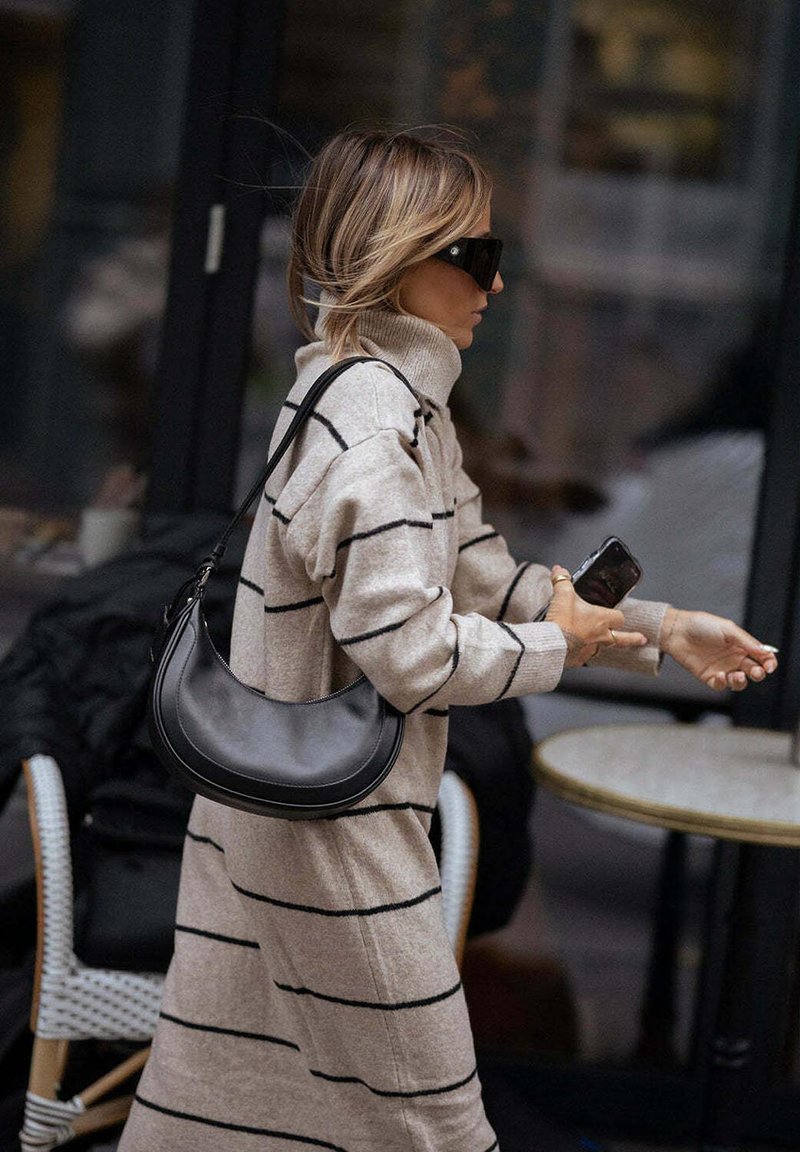Abito maglione beige con righe nere, colletto alto e vestibilità ampia; abbinato a una borsa a spalla nera. Accessorio: occhiali da sole neri oversize.