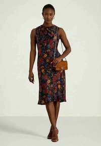 Γυναίκα με αμάνικο, μίνι φόρεμα σε σκούρο λουλουδάτο σχέδιο, κρατώντας μια μικρή καφέ τσάντα χιαστί, φορώντας καφέ παντόφλες με τακούνι, στέκεται μπροστά σε απλή φόντο.