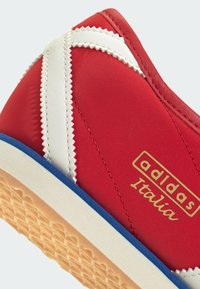 Baskets rouges Adidas Italia avec bande zigzag blanche, semelle en gomme, liseré bleu sur la semelle intermédiaire et texte doré Adidas Italia sur le côté.
