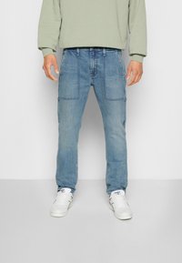 Světle modré denimové cargo kalhoty s dvěma předními kapsami a bočními kapsami. Nosič má na sobě šedý svetr a bílé tenisky. Rovný střih.