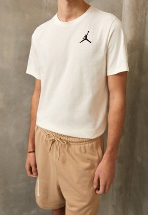 Mann trägt ein weißes Kurzarmhemd mit schwarzem Logo auf der Brust und beigen Shorts mit Kordelzug, steht an einer grauen Betonwand.