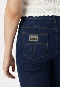 Mid-rise blue denim jeans featuring a grey logo patch on the back pocket, with orange stitching and a fitted design.