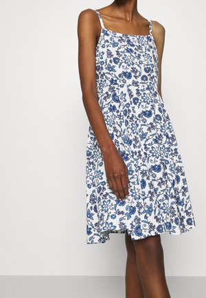 Mouwloze witte jurk met blauw bloemmotief, vierkante halslijn en dunne schouderbandjes, gedragen door een staand persoon tegen een effen achtergrond.