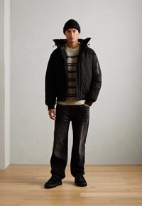 Black puffer jacket with zip pockets, striped cream sweater, dark jeans, and black sneakers. Model wears a black beanie. Light wooden floor.