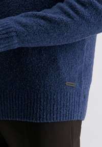 Blauer strukturierter Pullover mit geripptem Saum, der ein kleines schwarzes Markenlabel an der Seite aufweist, vor dunklen Hosen präsentiert.