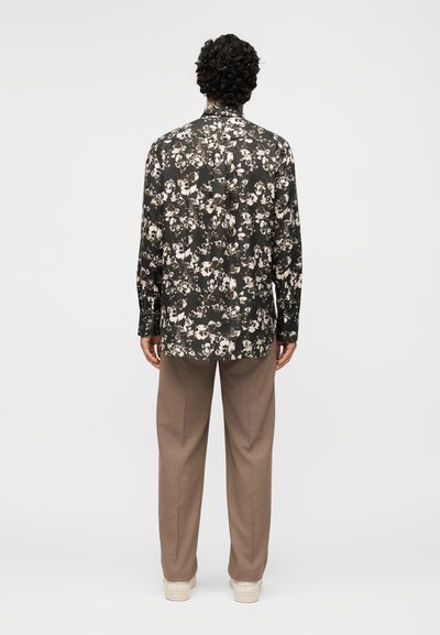 Άνδρας που στέκεται με την πλάτη γυρισμένη, φοράει μακρυμάνικο πουκάμισο με ασπρόμαυρα λουλούδια, μπεζ παντελόνι και λευκά αθλητικά παπούτσια σε απλό φόντο.