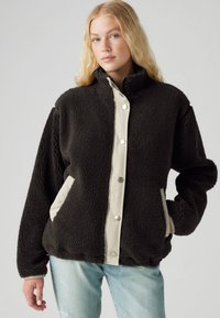 Chaqueta de forro polar negra con un cuello alto, botones crema y bolsillos laterales crema. Presenta una tela texturizada y un ajuste holgado.