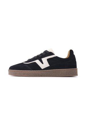 Zwarte suède sneaker met een dikke bruine zool, witte zijstreep, zwarte veters en een witte tong, zijaanzicht op witte achtergrond.