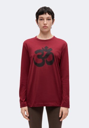 Joven con el pelo corto y oscuro que lleva una camiseta roja de manga larga con un gran símbolo Om negro en el frente.