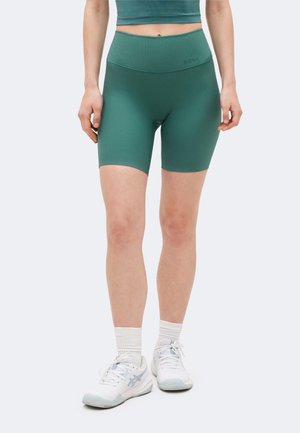 Nő zöld bordázott magasderekú kerékpáros rövidnadrágban, fehér sportzokniban és fehér futócipőben, egyszínű háttér előtt.