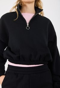 Μαύρο φούτερ με ψηλό λαιμό και μισό φερμουάρ, με ασημένιο τράβηγμα φερμουάρ και ελαστικό τελείωμα· φορεμένο πάνω από ένα ανοιχτό ροζ πουκάμισο.