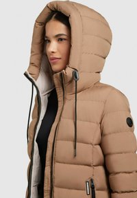 Beige puffer-takki, jossa on suuri pehmustettu huppu, etuvetoketju, joustavat nyörit ja musta logopainatus hihassa. Rakenne on teksturoitu.