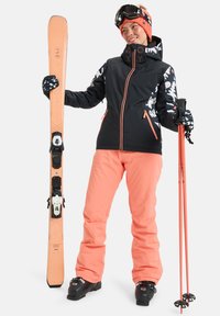 Conjunto de ski com uma jaqueta preta com detalhes em branco e coral, calças coral e botins de ski pretos. Segura um bastão de ski laranja e um ski.
