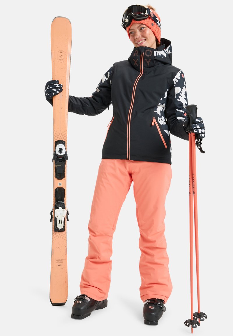 Conjunto de ski com uma jaqueta preta com detalhes em branco e coral, calças coral e botins de ski pretos. Segura um bastão de ski laranja e um ski.