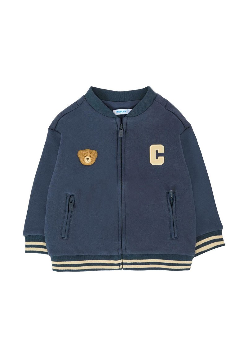 Giubbotto blu navy con zip realizzato in morbido tessuto. Presenta una toppa d'oro a forma di orso, una lettera "C" e polsini e orlo a righe. Due tasche con zip.