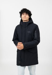 Schwarze Kapuzenparka-Jacke mit einer Reißverschlusstasche und mehreren Fronttaschen. Sie verfügt über einen glatten, strukturierten Stoff und ein Logo auf der Brust.