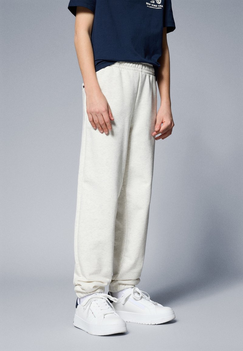 Personne portant un pantalon de survêtement couleur crème, des baskets blanches et un T-shirt bleu marine, debout devant un fond gris.