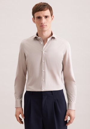 Man die een beige overhemd met knopen en donkere geplooide broek draagt, staand tegen een effen achtergrond.