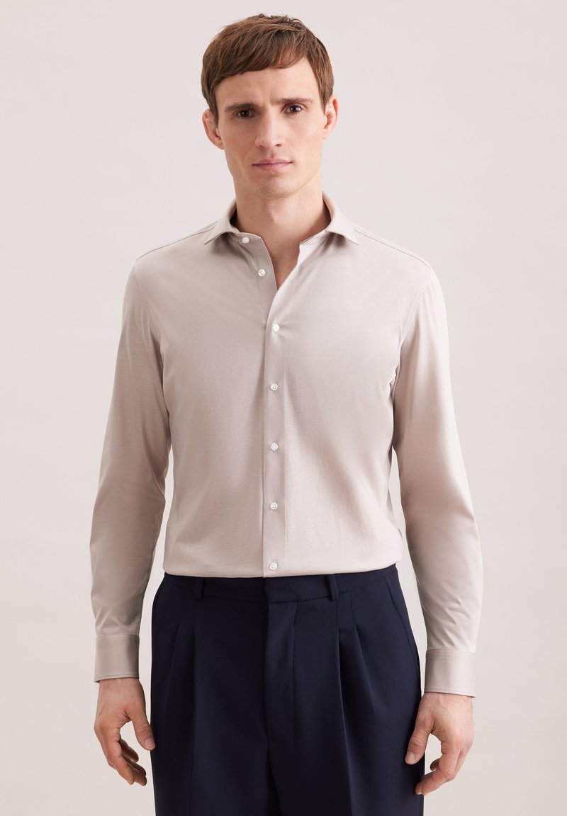 Homme portant une chemise beige boutonnée et un pantalon plissé foncé, debout devant un fond uni.