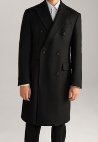 Cappotto in lana nero doppiopetto con rever a punta, due tasche frontali e bottoni neri, indossato sopra una camicia a righe blu chiaro e bianco.