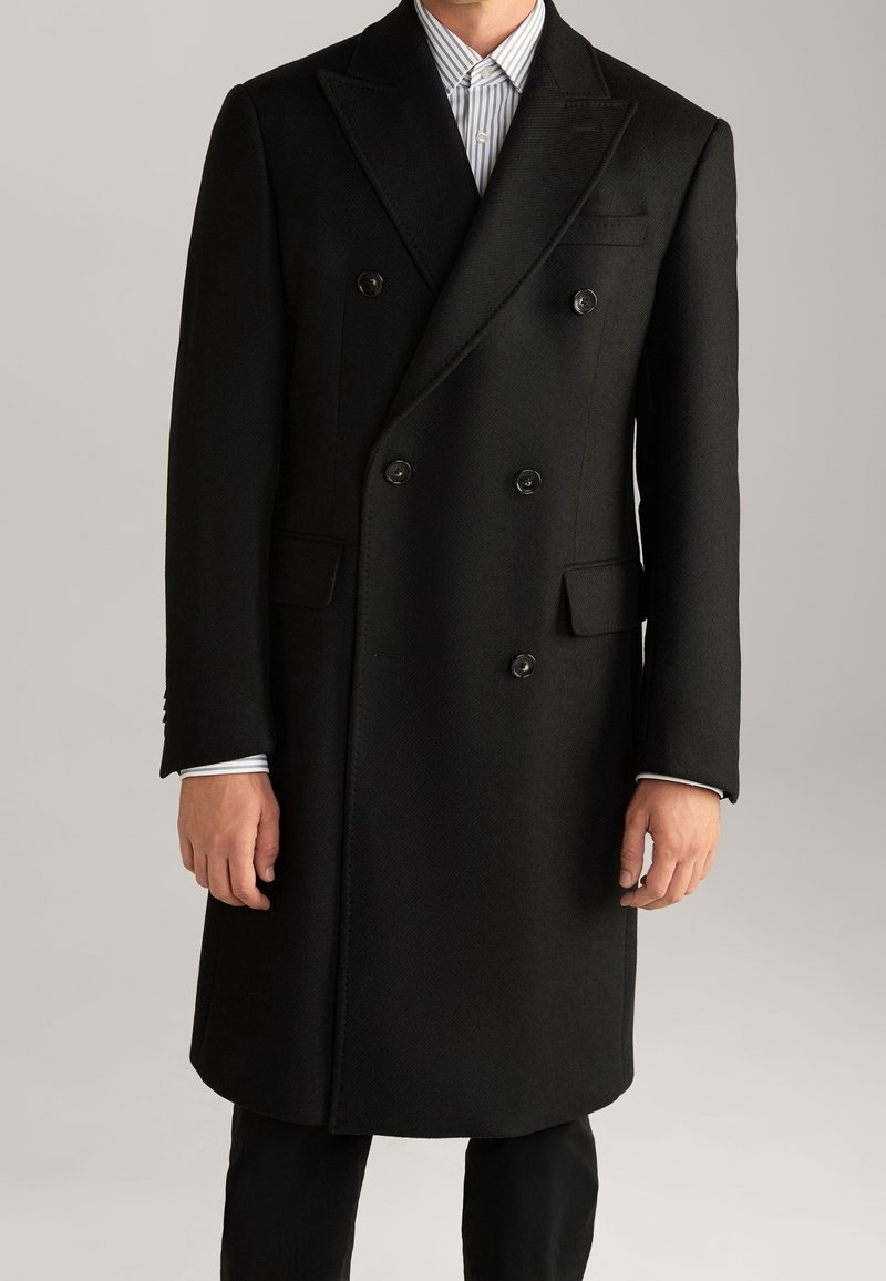 Cappotto in lana nero doppiopetto con rever a punta, due tasche frontali e bottoni neri, indossato sopra una camicia a righe blu chiaro e bianco.