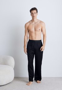 Black, lightweight pants with an elastic waistband, straight-leg design, worn by a model with a bare torso, standing on a light carpet.
