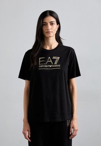 Crna pamučna majica s okruglim izrezom i kratkim rukavima. Sadrži zlatni EA7 Emporio Armani logo otisnut na prednjoj strani.