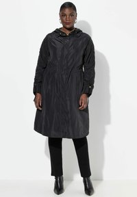 Svart, lättviktsjacka med huva och texturerade ärmar, åtdragen midja, dragkedja fram och sidofickor; i kombination med svarta byxor och klackade stövlar.