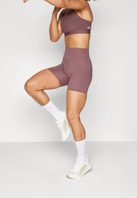 Fialová sportovní podprsenka a odpovídající vysoké kraťasy, vyrobené z elastického materiálu. Modelka má na sobě bílé ponožky a světlé sportovní boty.