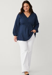 Navy blue long-sleeve blouse with a V-neck, textured fabric, front buttons, and a fitted waist; paired with white straight-leg trousers.