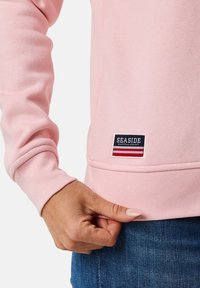 Felpa a maniche lunghe rosa chiaro con polsini a costine, caratterizzata da un patch logo "SEASIDE" blu navy con strisce rosse e bianche in fondo.