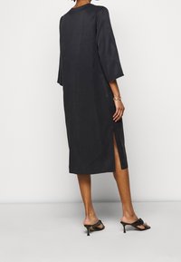 Mustan, polvipituisen mekon, jossa on 3/4-hihat, sivuaukot ja sileä pinta. Yhdistetty mustiin korkokenkiin, jotka esittelevät minimaalista muotoilua.