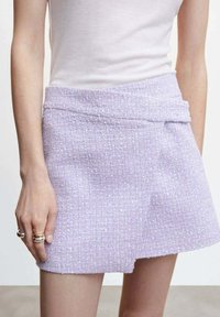 Mini-jupe en tweed lilas avec tissu texturé, taille haute et ourlet asymétrique, présentant un motif subtil et une finition lisse.