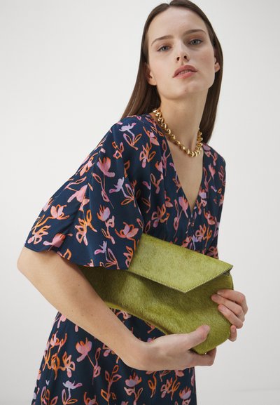 Grön kuvertväska med mjuk textur och form som ett kuvert, hålls av en person som bär en klänning med blommönster och ett kraftigt guldhalsband.