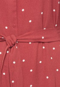 Červené šaty s bílými puntíky, vyrobené z texturované látky, s knoflíkovým zapínáním a opaskem se zavázáním v pase.