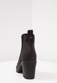 Czarny botek na obcasie z teksturowaną cholewką z tkaniny, elastycznymi panelami po bokach oraz pętelką do zakładania; posiada masywną, żłobioną podeszwę.