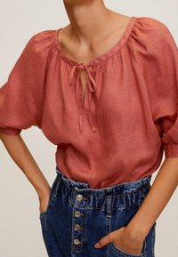 Blouse en lin couleur rouille avec un col rond, détail à nouer et manches courtes bouffantes, associée à un short en jean taille haute avec boutons.