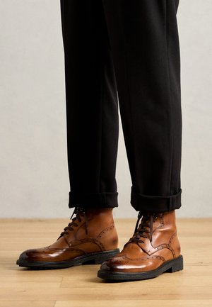 Bruine leren enkellaarzen met brogue-details, zwarte rubberen zolen en zwarte veters, gecombineerd met opgerolde zwarte broeken op een houten vloer.