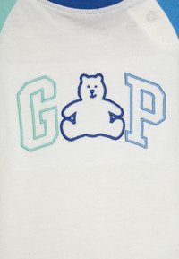 Hvit bomullsskjorte med blå og grønne raglanermer. Den har en brodert bjørn og bokstavene 'G', 'P' i blå og grønne nyanser.