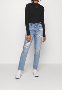 Černý dlouhý rukáv s žebrovanou strukturou v kombinaci s světle modrými roztrhanými džínami a bílými teniskami, které předvádějí uvolněný střih a detaily s distresovaným denimem.