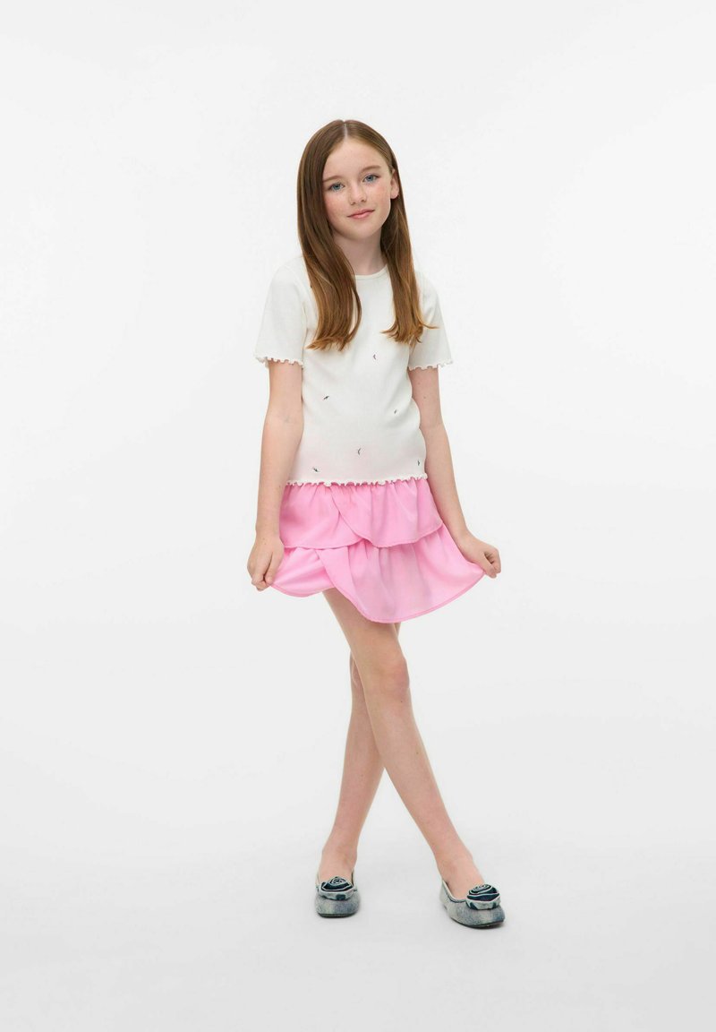 Top blanco de manga corta con ribetes ondulados, combinado con una falda rosa de volantes en capas. Presenta zapatos grises con acentos decorativos.