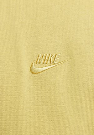 Gelbes Material mit glatter Textur zeigt ein gesticktes Nike-Logo in einem etwas dunkleren Gelb. Einfaches Design und klare Linien.