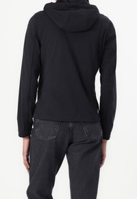 Svart huva jacka med en slät, matt yta, med långa ärmar och en kortad skärning. Bärs över mörka denimjeans med märkbara sömmar.