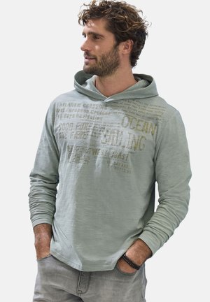 Man with curly hair wearing a light green hoodie with sailing-themed text, standing with hands in pockets, looking to his left.