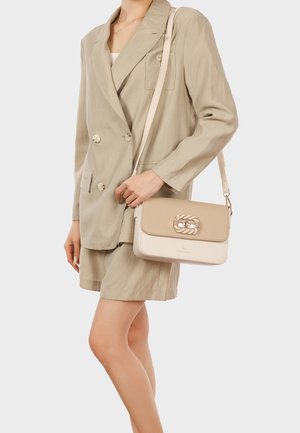 Donna che indossa un blazer beige e pantaloncini, con una borsa a tracolla beige e crema con fibbia dorata sulla spalla.