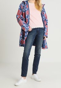 Waterdichte bloemenregenjas in het blauw met rode bloemen, gelaagd over een lichtgestreept shirt. Donkere jeans en witte sneakers maken de outfit compleet.