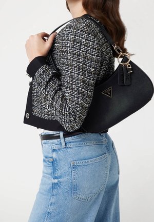 Femme portant une veste en tweed noir et blanc et un jean bleu clair, tenant un sac à bandoulière noir Guess avec des ferrures dorées sur l'épaule.