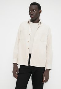 Bézs színű, texturált, gombos ing két mellzsebbel és arany gombokkal; világosszürke felsőre viselve, fekete nadrággal párosítva.