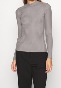 Graues, geripptes Langarmoberteil mit hohem Kragen, das ein enganliegendes Design und eine glatte Textur aufweist, kombiniert mit schwarzen Hosen.