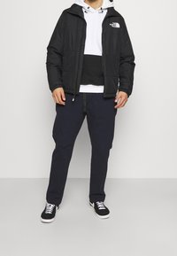 Černá zateplená bunda přes bílou a černou mikinu, spárovaná s námořnickými kalhotami a černými teniskami. Obsahuje akcenty loga a stahovací kapuci.